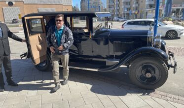 Mies seisoo mustan vintage-tyylisen auton edessä aurinkoisena päivänä kaupunkimaisemassa. Hänellä on päällään sininen paita ja kuviollinen takki. Auton ovi on auki ja taustalla näkyy modernia kaupunkirakennusta ja parkkeerattuja autoja.